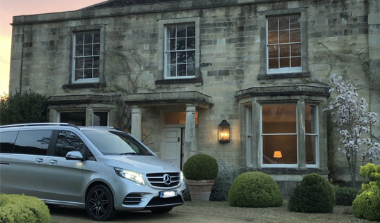 Silver Mercedes van parked in front of a stone house with multiple windows.